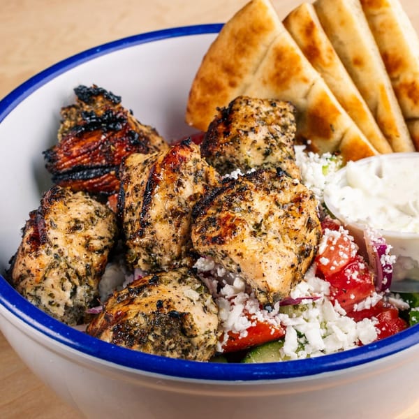 Grilled chicken skewers with feta, tomatoes, cucumbers, and pita bread in a white bowl with a blue rim.