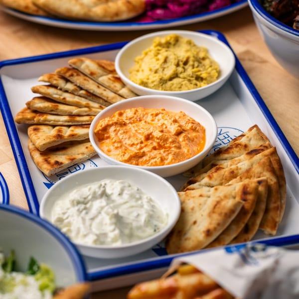 A white tray with blue trim holds pita bread and three bowls of dips: hummus, red pepper, and tzatziki.