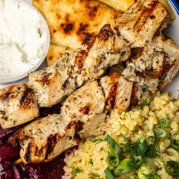 Grilled chicken skewers with rice, red cabbage, pita bread, and a side of tzatziki sauce.