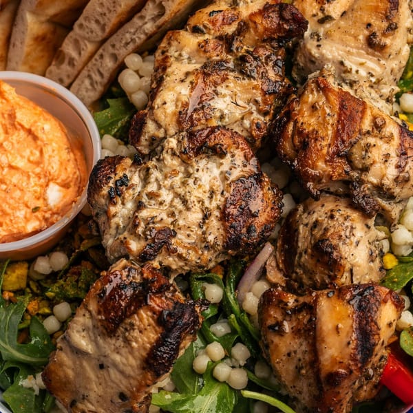 Grilled chicken pieces with a side of red dip, bread, and a salad with white beans.