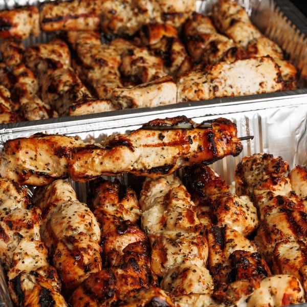 Close-up of grilled chicken skewers and pieces in two foil trays, seasoned with herbs.