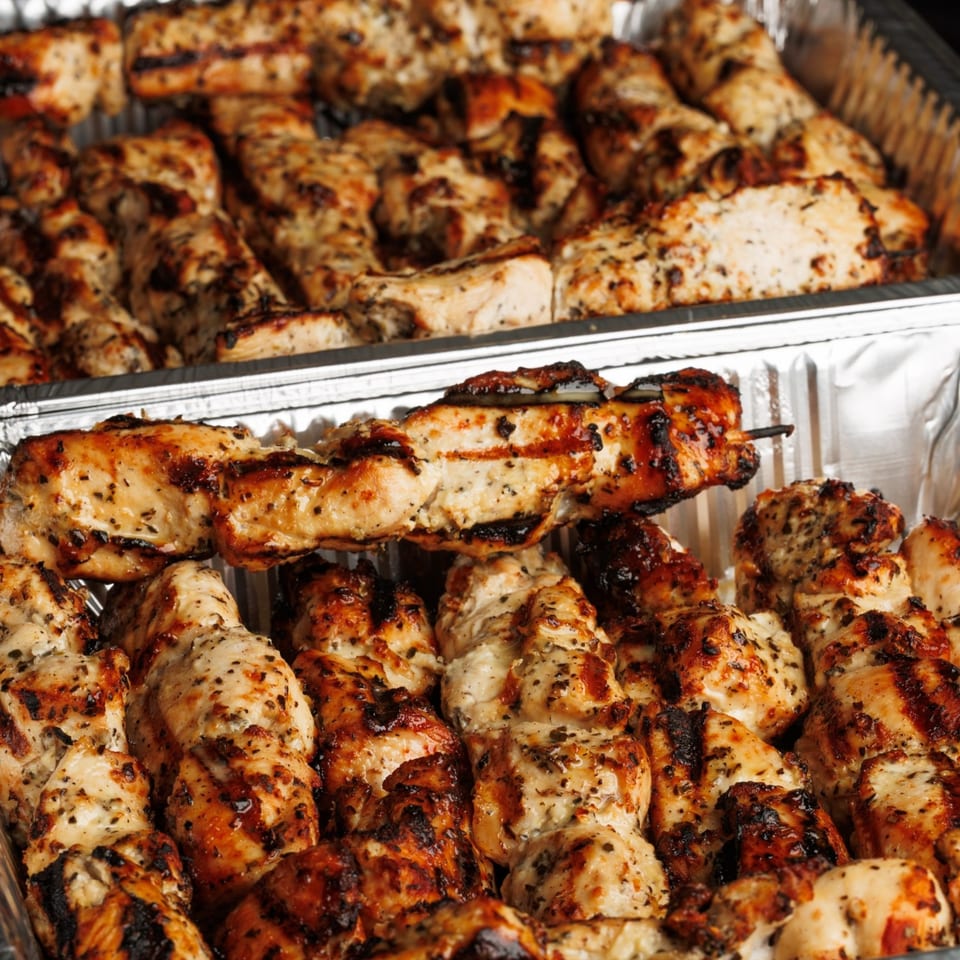Close-up of grilled chicken skewers and pieces in two foil trays, seasoned with herbs.